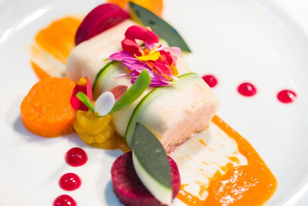 Ein Gourmetgericht mit pochiertem Fisch, umhüllt von Zucchini und garniert mit bunten essbaren Blüten. Dazu gibt es Rote-Bete und Karottenpüree sowie rote Soßentupfen.