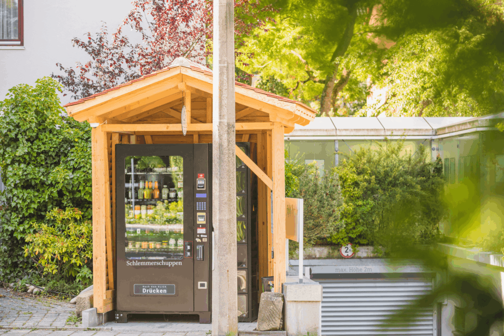 Ein kleiner Holzunterstand mit einem Lebensmittel-Automaten steht an einer sonnigen Straße. Umgeben von üppigem Grün schafft er eine friedliche, einladende Atmosphäre vor dem nachhaltigen Hotel Luise.
