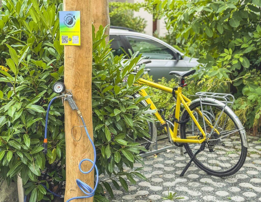 Ein gelbes Fahrrad steht auf einem Kopfsteinpflasterweg neben dem nachhaltigen Hotel Luise. An einem Holzpfosten sind eine Reifenpumpe und ein Manometer befestigt. Ein graues Auto ist teilweise zu sehen.