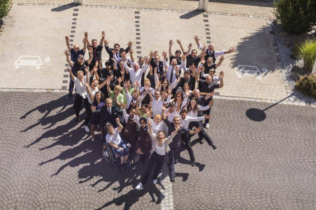 Eine bunte Gruppe von Menschen aus dem SCHWARZWALD PANORAMA-Team steht draußen auf einem gepflasterten Platz, lächelt und reckt freudig die Hände in die Höhe. Die Szene vermittelt ein Gefühl von Zusammengehörigkeit und Feierlichkeit.