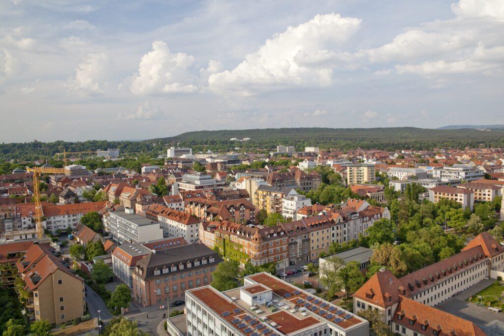 Luftaufnahme der malerischen Stadt Erlangen in Bayern mit ihren Gebäuden mit roten Dächern, üppig grünen Bäumen und einer Kulisse aus sanften Hügeln unter einem teilweise bewölkten blauen Himmel.