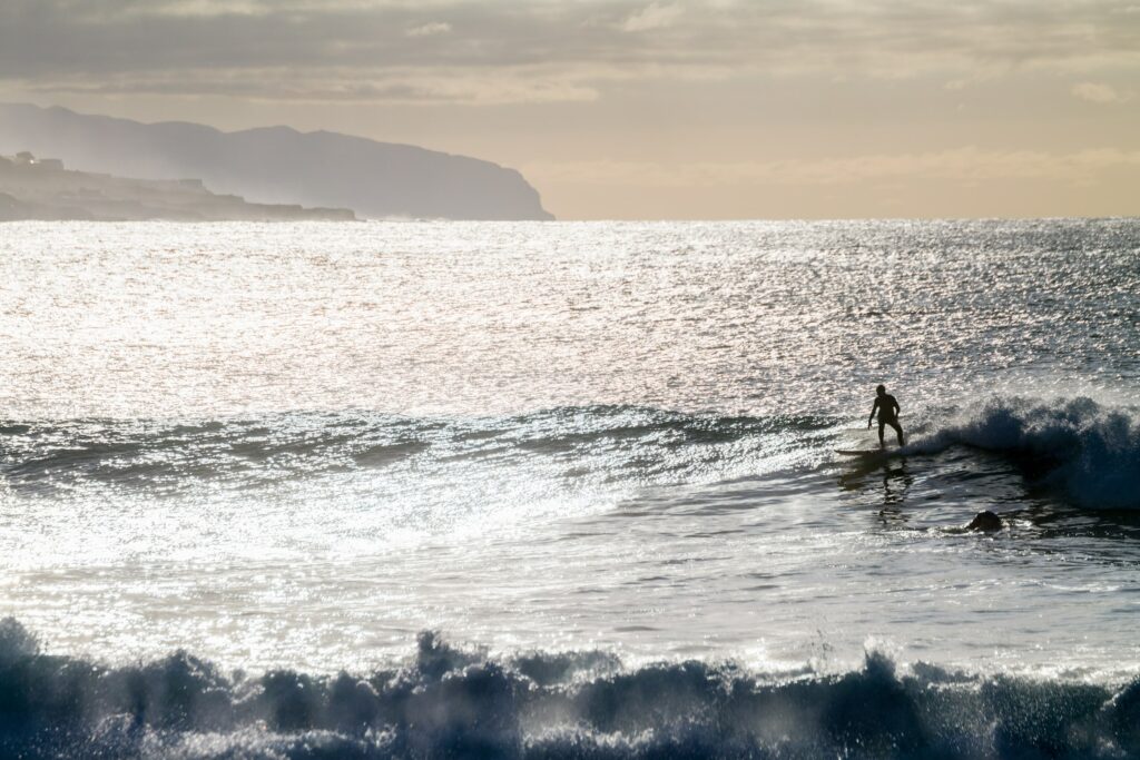 Ein Surfer reitet auf einer sanften Welle auf dem glitzernden Atlantik in der Nähe des nachhaltigen Wellnesshotels OCÉANO unter einem bewölkten Himmel. In der Ferne zeichnen sich die Klippen des Anaga-Gebirges am Horizont ab und vermitteln eine ruhige und abenteuerliche Stimmung.