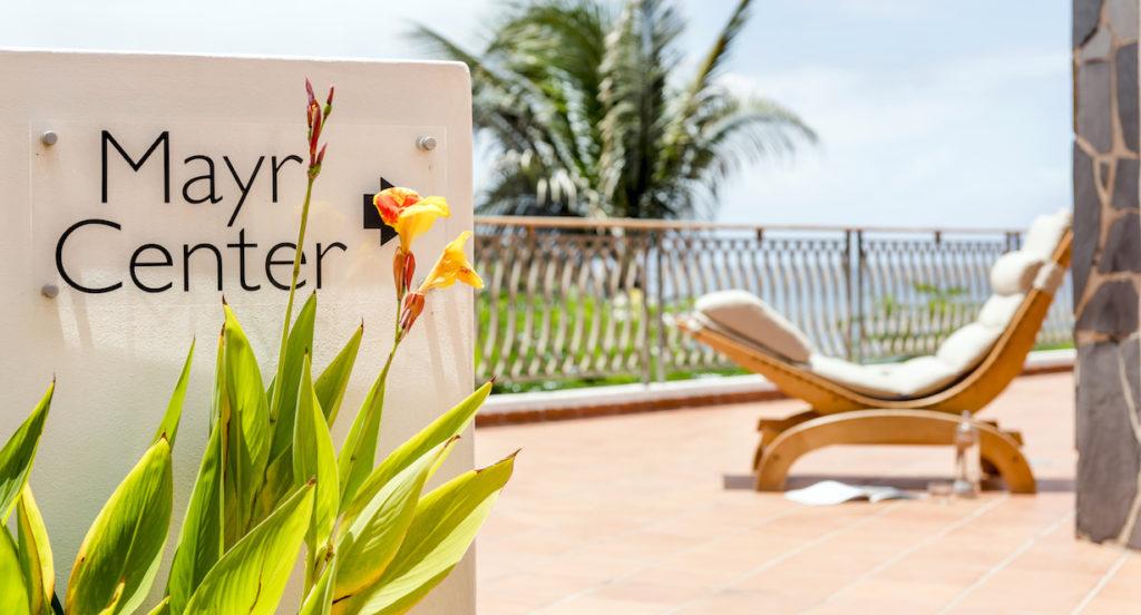 Schild mit der Aufschrift „Mayr Center“ neben leuchtend gelben Blumen. Im Hintergrund ein gepolsterter Liegestuhl auf einer Terrasse mit Palmen, der eine ruhige, entspannte Atmosphäre schafft.