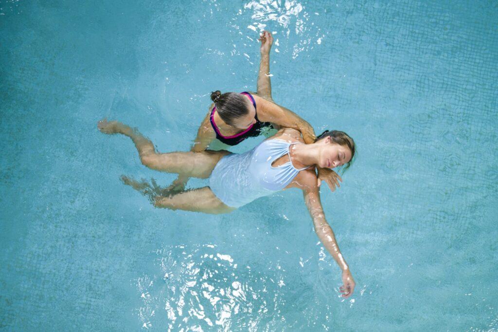 Zwei Frauen befinden sich in einem Thalasso-Pool im nachhaltigen OCÉANO Health Spa Hotel. Eine Frau in einem schwarzen Badeanzug stützt die andere, die entspannt in einem weißen Badeanzug auf dem Rücken treibt.