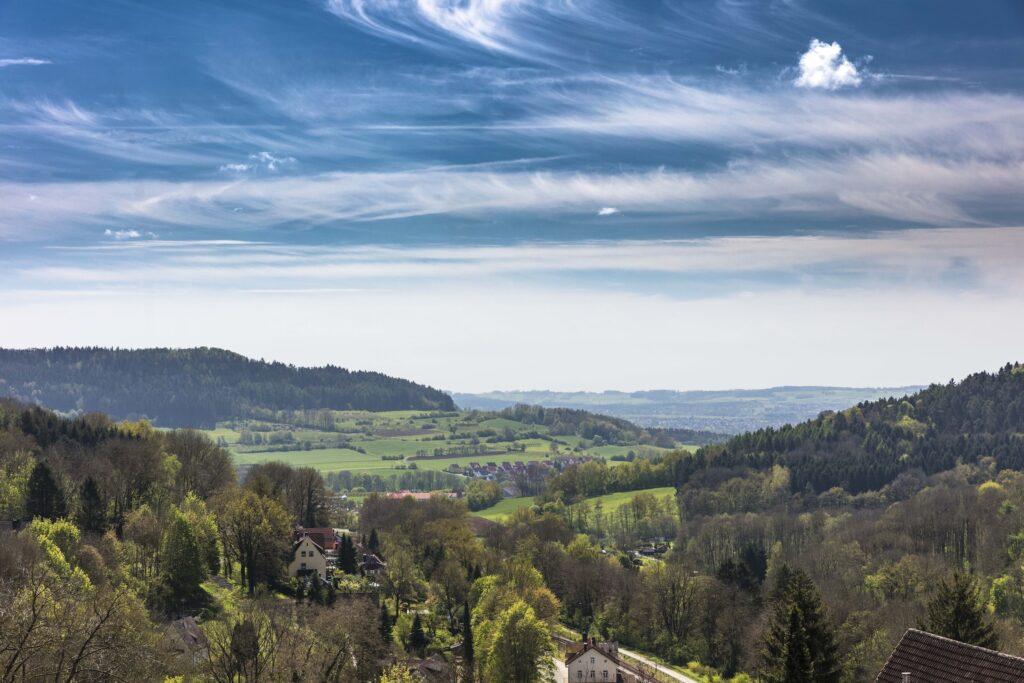 Malerischer Blick auf ein üppiges Tal bei Erlangen in Franken mit sanften grünen Hügeln und verstreuten Häusern unter blauem Himmel mit dünnen Wolken, der Ruhe und Weite vermittelt.