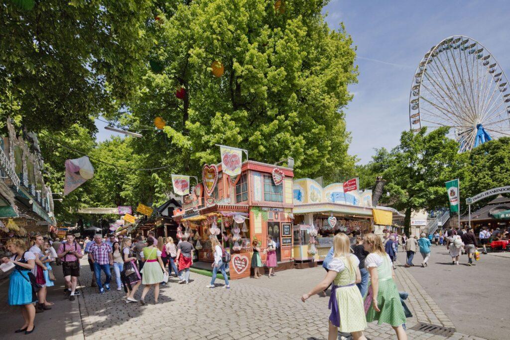 Menschen in traditioneller Tracht genießen die lebhafte Bergweihkirch in Erlangen mit bunten Ständen unter großen Bäumen und einem Riesenrad im Hintergrund, das für eine festliche Atmosphäre sorgt.