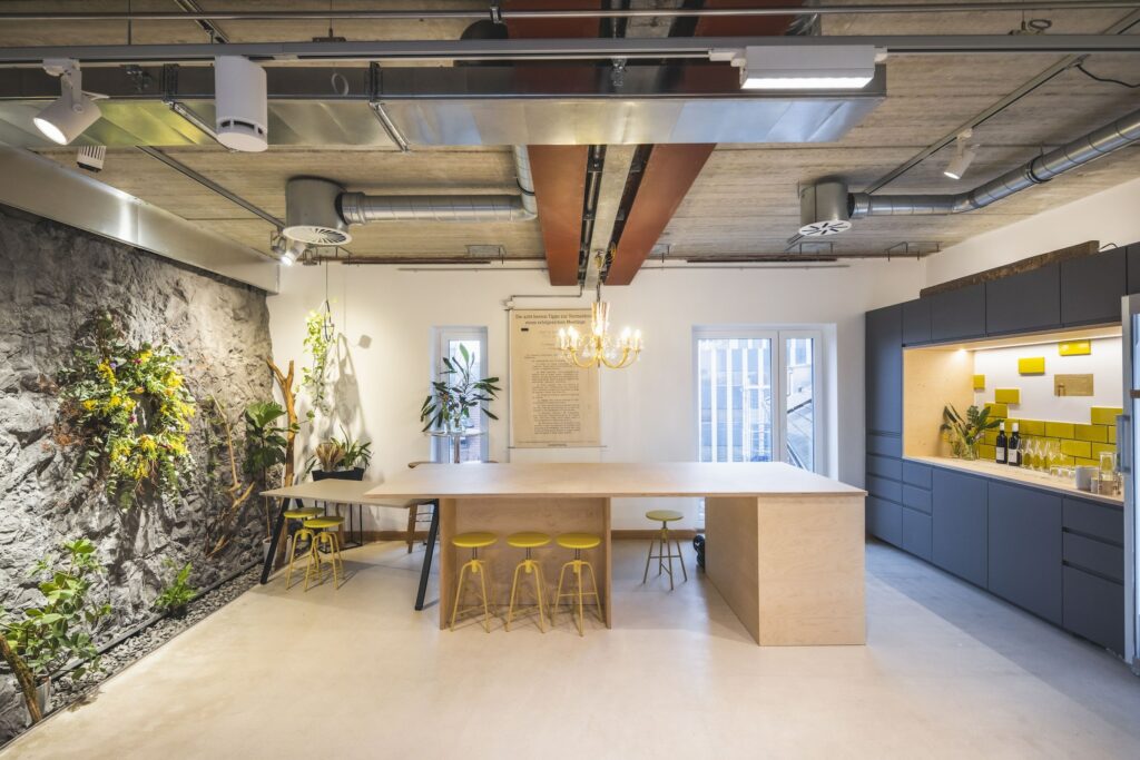 Der moderne Businessraum im nachhaltigen Hotel Luise mit industriellem Design verfügt über eine lange Holzinsel, gelbe Hocker und graue Schränke. Eine Felswand mit Pflanzen sorgt für Grün und Wärme.