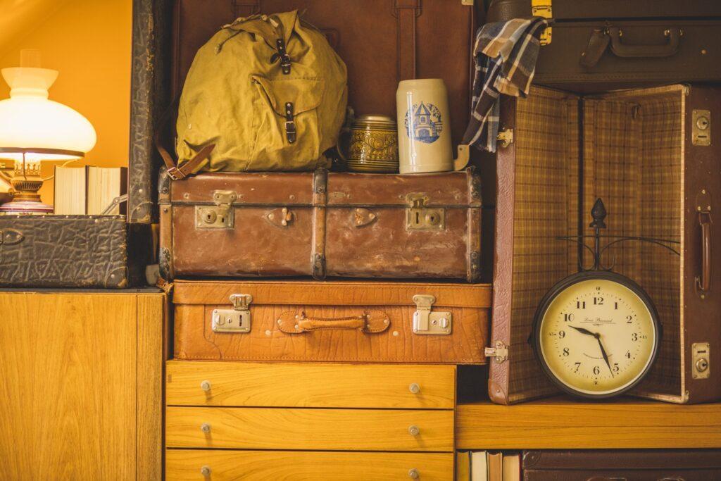 Vintage-Themenzimmer im nachhaltigen Hotel Luise mit gestapelten braunen Lederkoffern, einem gelben Rucksack, Büchern und einem offenen Koffer mit Karomuster, in dem eine altmodische Uhr steht. Warme Beleuchtung sorgt für eine nostalgische Atmosphäre.