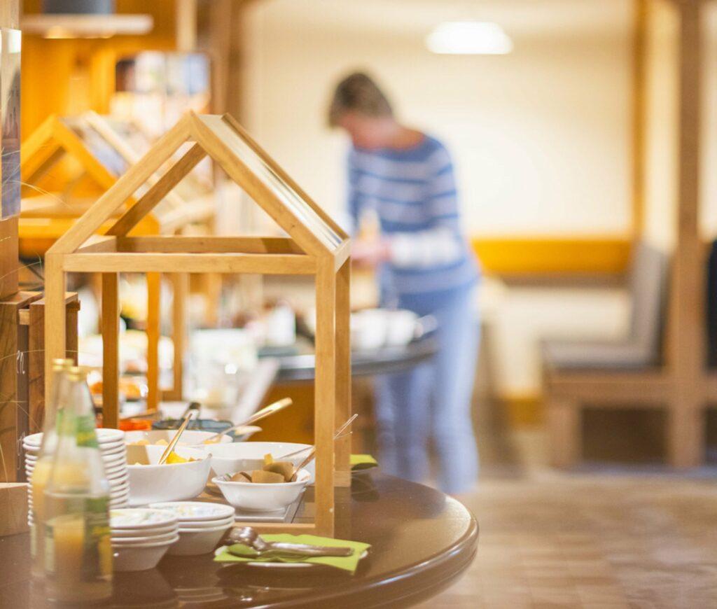 Zero-Waste-Buffet im nachhaltigen Hotel Luise mit verschiedenen Schüsseln und Tellern unter einem Holzrahmen, unscharf abgebildete Person in gestreiftem Hemd, die eine ungezwungene, einladende Atmosphäre schafft.