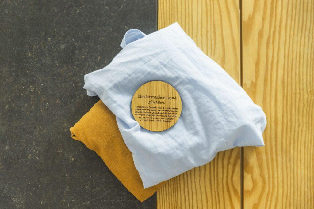 Hellblauer und senffarbener Stoff sind ordentlich auf einer dunklen Holzoberfläche gefaltet. Darauf liegt ein rundes Holzschild mit einer Aufschrift, die Wärme und Schlichtheit vermittelt. Green Project im Eco Hotel Luise
