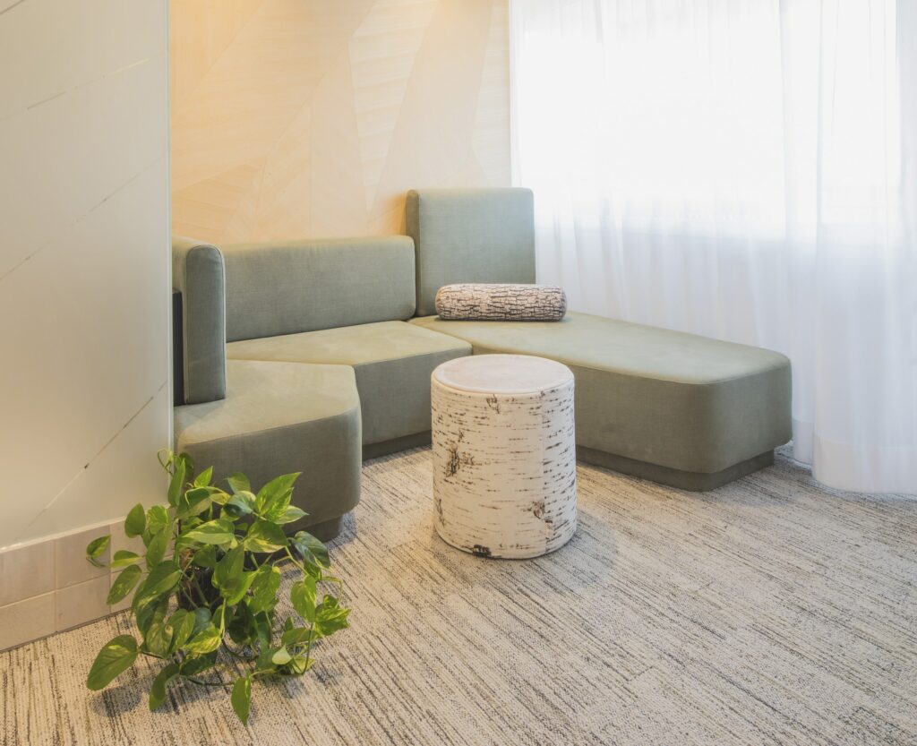 Moderne Wohnzimmerecke im nachhaltigen Hotel Luise mit einem hellgrünen L-förmigen Sofa, einem runden Hocker mit Birkenmuster, einer grünen Zimmerpflanze und sanftem Tageslicht, das durch transparente Vorhänge fällt.