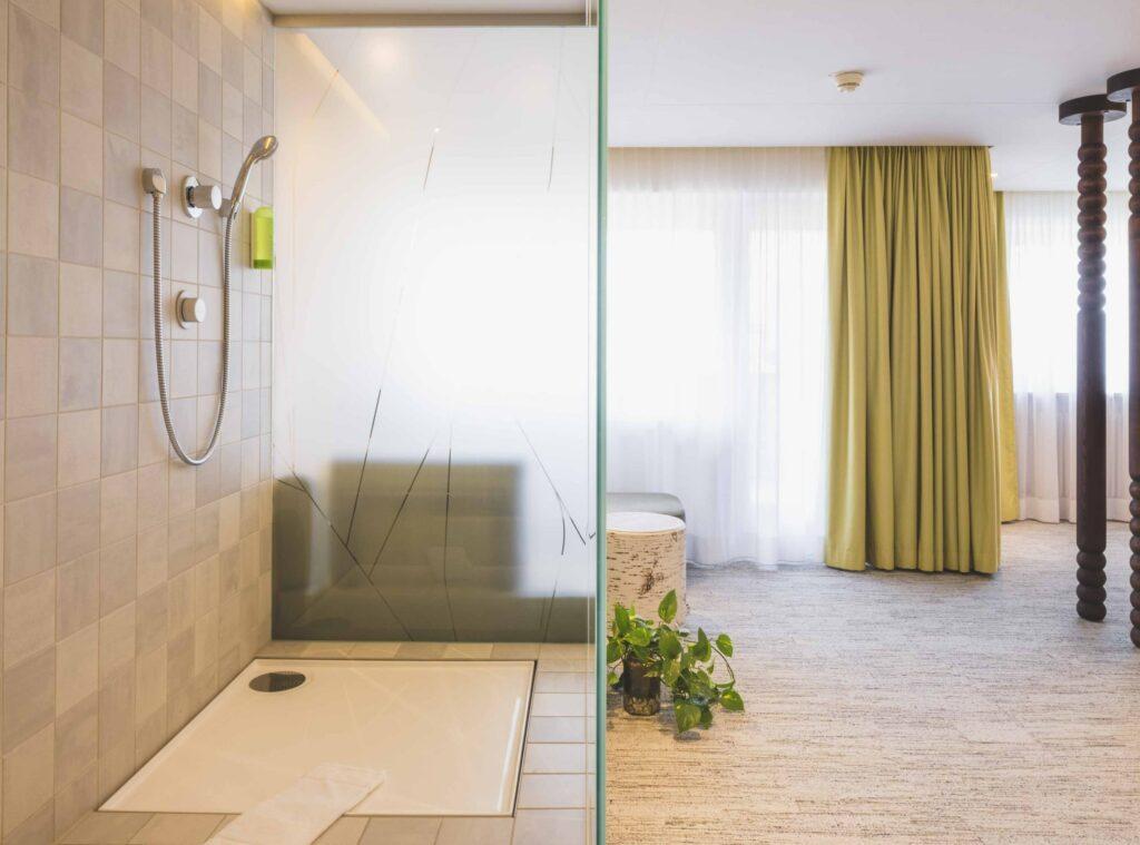 Minimalistisches Zimmer im Öko-Hotel Luise mit Duschbereich auf der linken Seite, gefliesten Wänden und einer Handbrause. Auf der rechten Seite befindet sich ein heller Raum mit grünen Vorhängen, einer Pflanze und einem Holzhocker.
