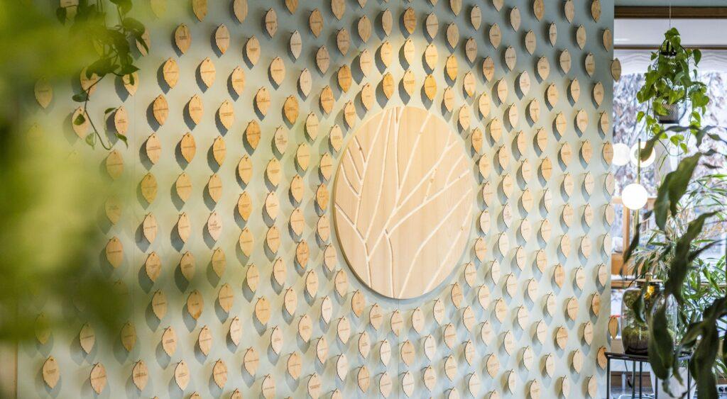 Das grüne Projekt des wegweisenden Hotels Luise: Die „Wall of Change“ mit einem kreisförmigen Kunstwerk umgeben von blattförmigen Holztafeln. In der Nähe hängen Grünpflanzen, die eine beruhigende Atmosphäre schaffen.
