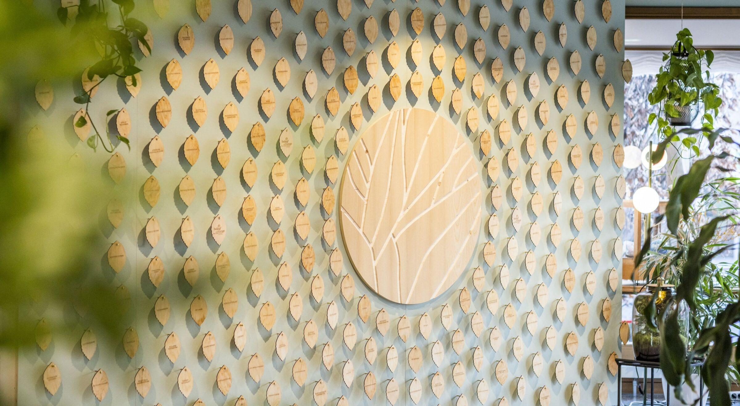 Das grüne Projekt des wegweisenden Hotels Luise: Die „Wall of Change“ mit einem kreisförmigen Kunstwerk umgeben von blattförmigen Holztafeln. In der Nähe hängen Grünpflanzen, die eine beruhigende Atmosphäre schaffen.