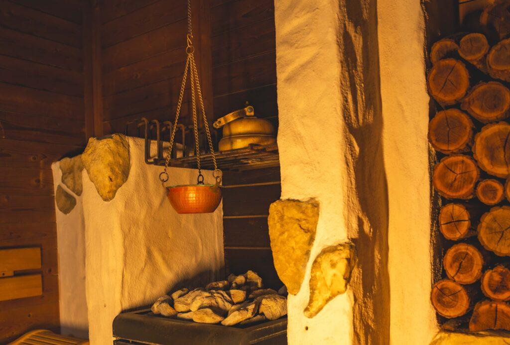 Gemütliches Sauna-Interieur mit warmer Beleuchtung im Spa des nachhaltigen Hotels Luise in Erlangen. Mit gestapeltem Brennholz, großen Steinen, einem Messingkessel und einem hängenden Kupfertopf, die eine rustikale, ruhige Atmosphäre schaffen.