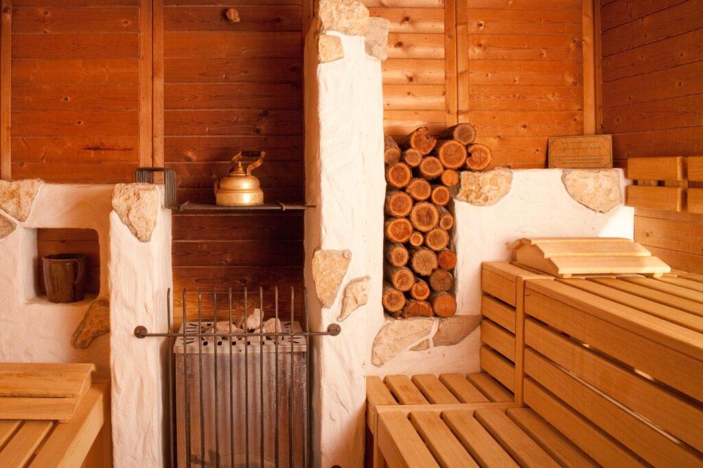 Gemütliche Sauna im nachhaltigen Hotel Luise in Erlangen mit Holzbänken, einer gestapelten Holzscheitwand und einem rustikalen Ofen mit einem Wasserkessel. Warme, einladende Atmosphäre mit holzgetäfelten Wänden.