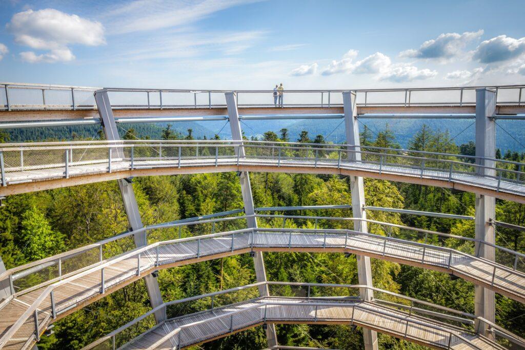 Ein spiralförmiger Aussichtssteg ragt über den üppig grünen Schwarzwald empor und führt zu einer Plattform, auf der zwei Menschen unter strahlend blauem Himmel die weitläufige Aussicht genießen.