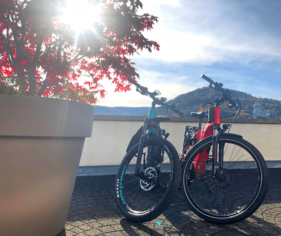 Zwei E-Bikes lehnen unter einem klaren Himmel an einer Steinmauer, eingerahmt von einer strahlenden Sonne hinter roten Herbstblättern. Im Hintergrund ein ruhiger Bergblick über den Schwarzwald.