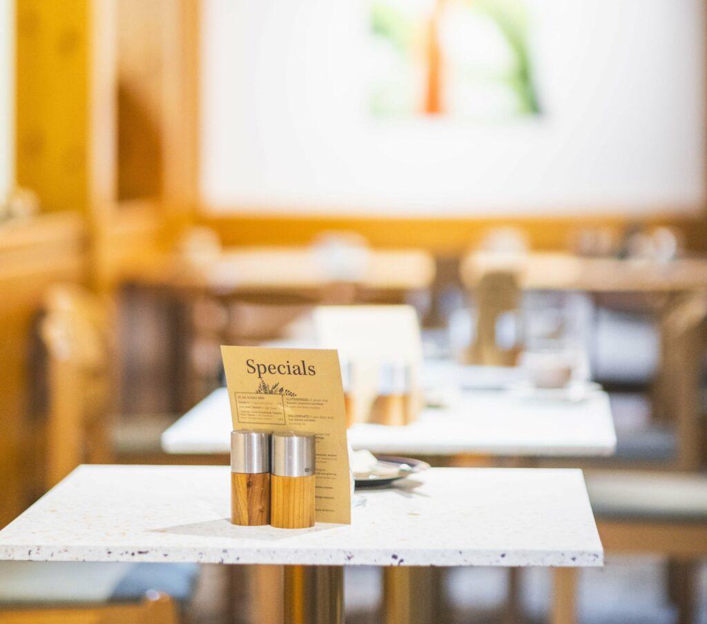 Helles Restaurantinterieur des Hotels Luise mit Fokus auf einem weißen Tisch (aus recycelten Kühlschränken) mit einer Tageskarte und Salz- und Pfefferstreuern aus Holz. Unscharfer Hintergrund mit Tischen.