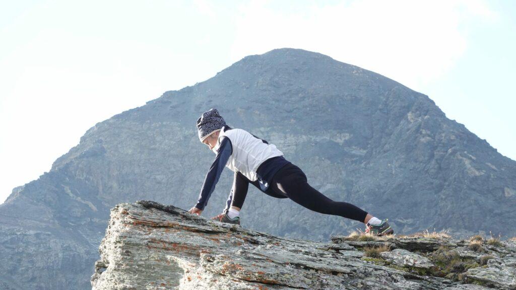 Eine Person in Sportbekleidung macht auf einem Felsvorsprung vor einer Bergkulisse Dehnübungen. Die Szene vermittelt Entschlossenheit und Ruhe.