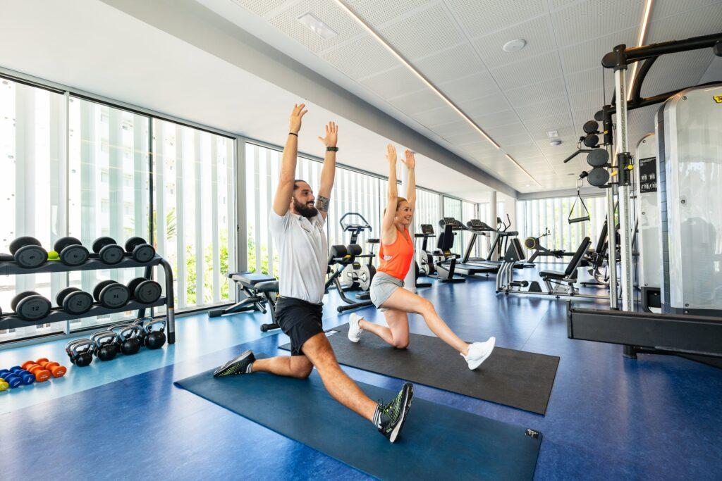 Ein Mann und eine Frau führen in einer hellen Sporthalle Ausfallschritte mit erhobenen Armen auf Matten aus. Hanteln und Kettlebells sind sichtbar und erzeugen einen energischen und konzentrierten Ton. Bewegungsprogramm ist Teil der Regeneration im Wellnesshotel OCÉANO auf Teneriffa