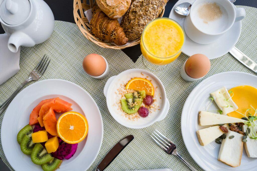 Ein reichhaltiges Frühstücksbuffet mit bunten Früchten auf einem Teller, Käse und Beilagen, zwei gekochten Eiern, Joghurt mit Früchten, einem Korb mit Gebäck, Orangensaft und einer Tasse Kaffee – all das im nachhaltigen Restaurant La Marea des OCÉANO Health Spa Hotels auf Teneriffa.