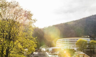 Sonnenbeschienene Szene des modernen, runden Gebäudes des nachhaltigen Hotels SCHWARZWALD PANORAMA inmitten von Dächern und Bäumen. Das Sonnenlicht strahlt und unterstreicht die ruhige Bergkulisse mit einem ruhigen, warmen Ton.