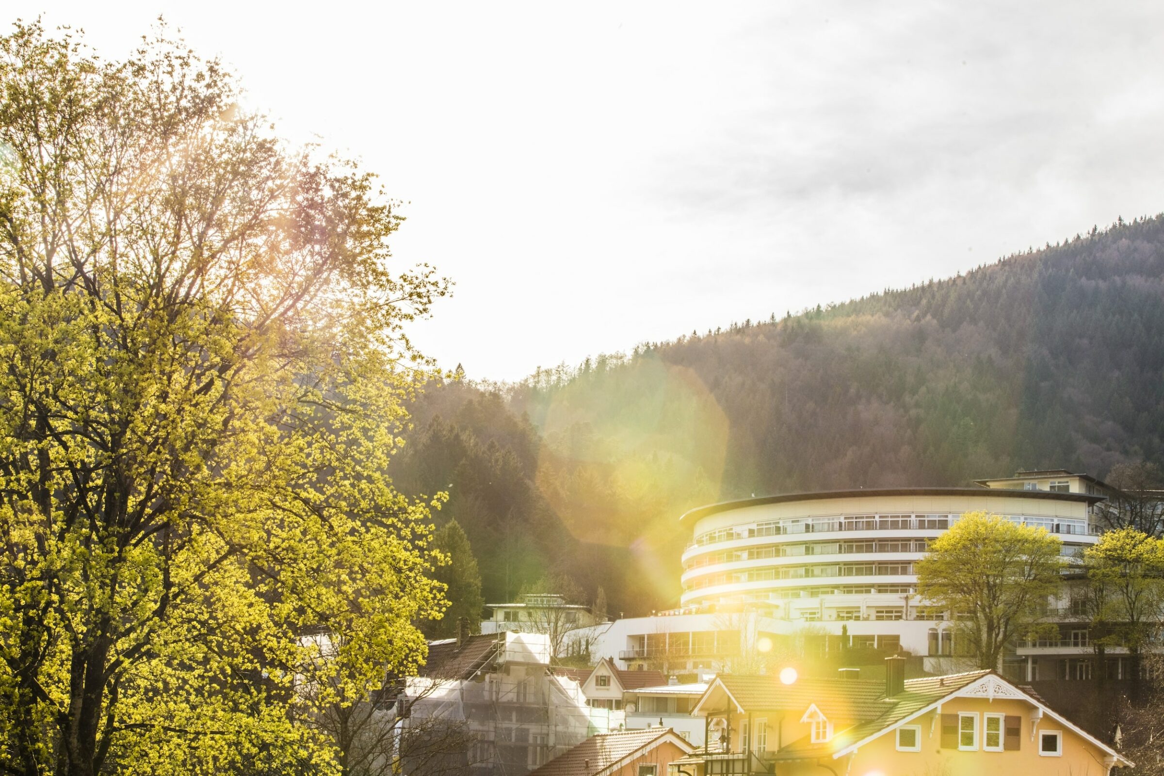 Sonnenbeschienene Szene des modernen, runden Gebäudes des nachhaltigen Hotels SCHWARZWALD PANORAMA inmitten von Dächern und Bäumen. Das Sonnenlicht strahlt und unterstreicht die ruhige Bergkulisse mit einem ruhigen, warmen Ton.