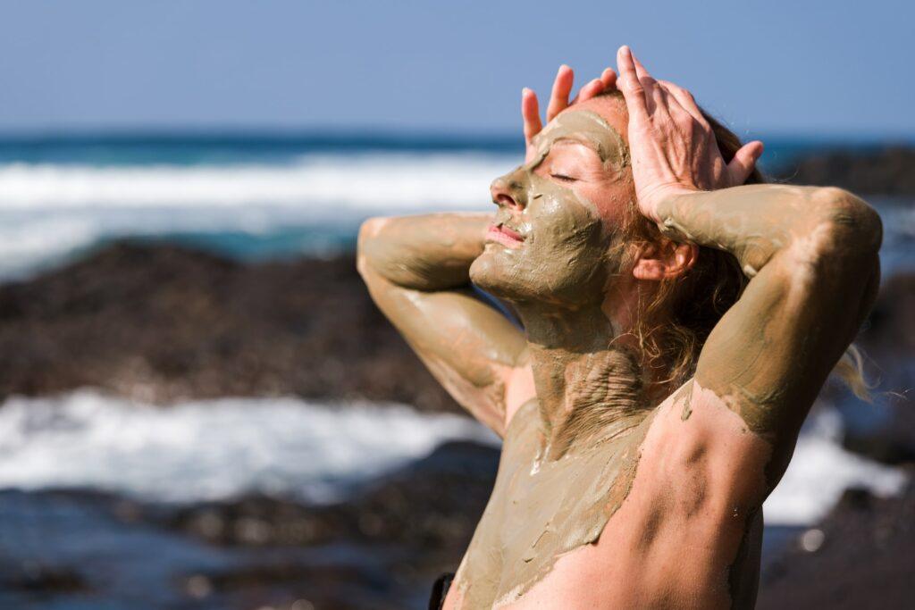 Eine Frau genießt einen sonnigen Strand, ihr Gesicht und ihr Körper sind mit grünem Meeresschlamm bedeckt. Sie hat die Augen geschlossen und die Hände auf dem Kopf, sie strahlt Entspannung und Zufriedenheit aus – Das Ergebnis eines Aufenthalts im OCÉANO Health Spa Hotel