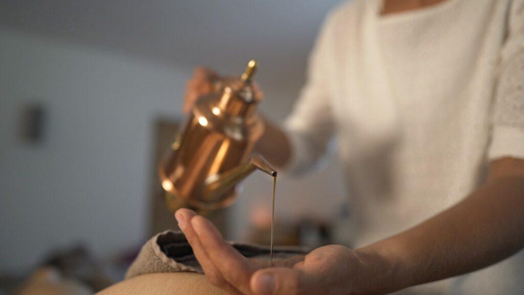 Eine Person in einem weißen Oberteil gießt Öl aus einem Kupfertopf auf ihre Handfläche für eine Ayurveda-Ölmassage im veganen Hotel LA VIMEA und schafft so eine ruhige und friedliche Atmosphäre. Der Hintergrund ist sanft unscharf.