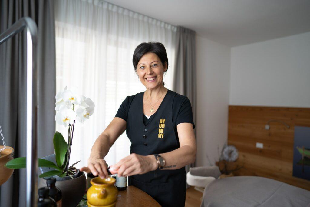 Lächelnde Frau in der Spa-Umgebung des ganzheitlich veganen Hotels LA VIMEA, bekleidet mit einer schwarzen Uniform mit goldener Beschriftung. Sie steht neben einem Tisch mit einer weißen Orchidee und trägt Creme aus einem kleinen Tiegel auf.