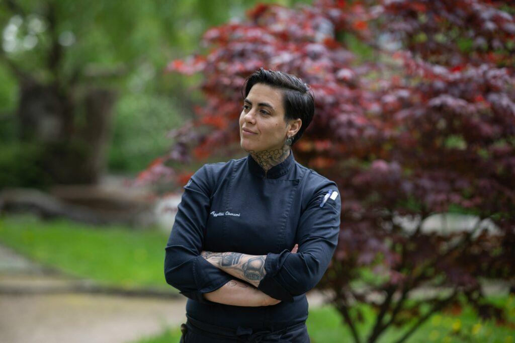 Die vegane Köchin Aggeliki Charami in dunkler Uniform steht selbstbewusst mit verschränkten Armen vor einem Hintergrund aus üppigem Grün und leuchtend rotem Laub und strahlt Stolz und Professionalität aus.