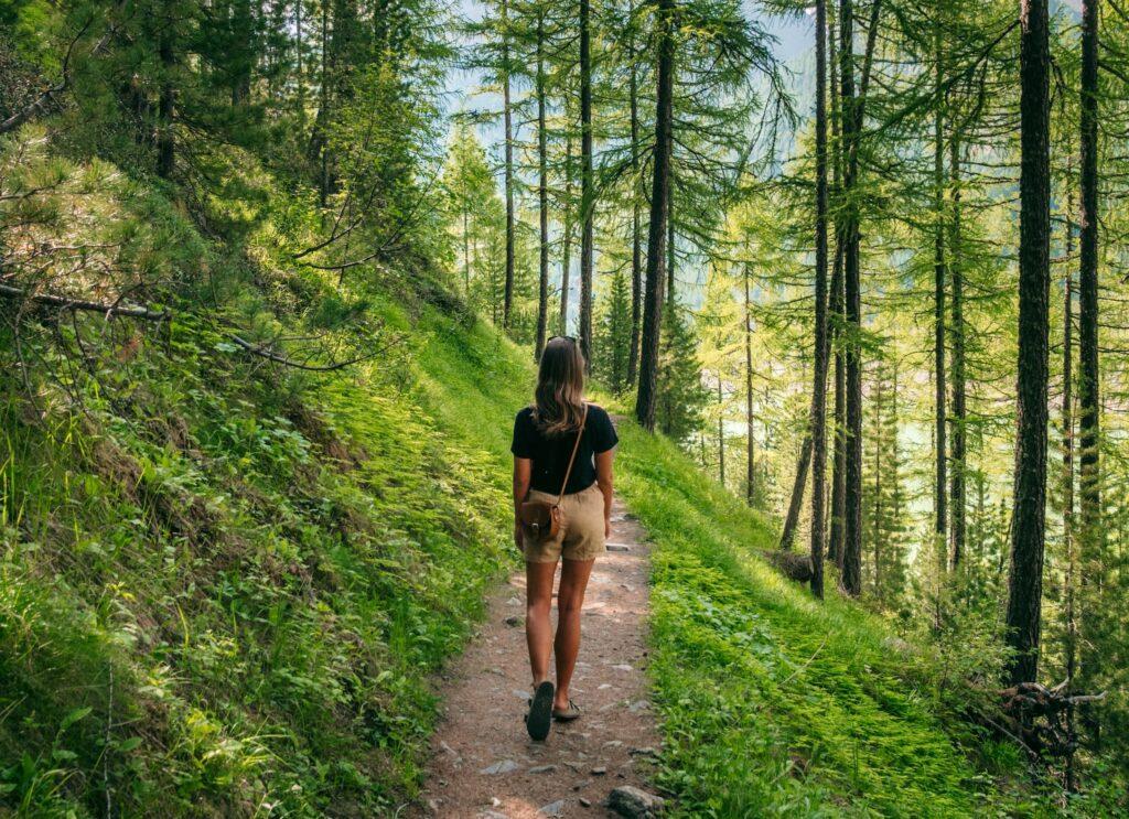 Eine Frau in Freizeitkleidung spaziert durch einen üppigen, sonnenbeschienenen Waldweg in Südtirol in der Nähe von Naturns, umgeben von hohen Bäumen und viel Grün, was ein Gefühl von Ruhe und Abenteuer hervorruft.
