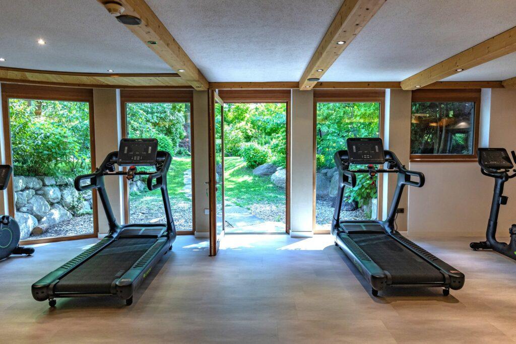 Der Fitnessraum des veganen Hotels LA VIMEA verfügt über moderne Laufbänder mit Blick auf große Fenster, die einen ruhigen Garten mit üppigem Grün und Felsen zeigen. Die Szenerie wirkt friedlich und einladend.