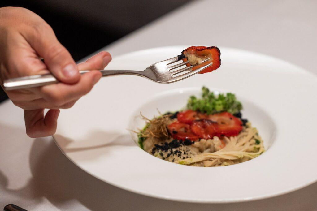 Eine Hand hält eine Gabel mit einer gegrillten Erdbeerscheibe über einem weißen Teller mit einem veganen Gourmetgericht, darunter Pasta und Kräuter, auf einer weißen Tischdecke.