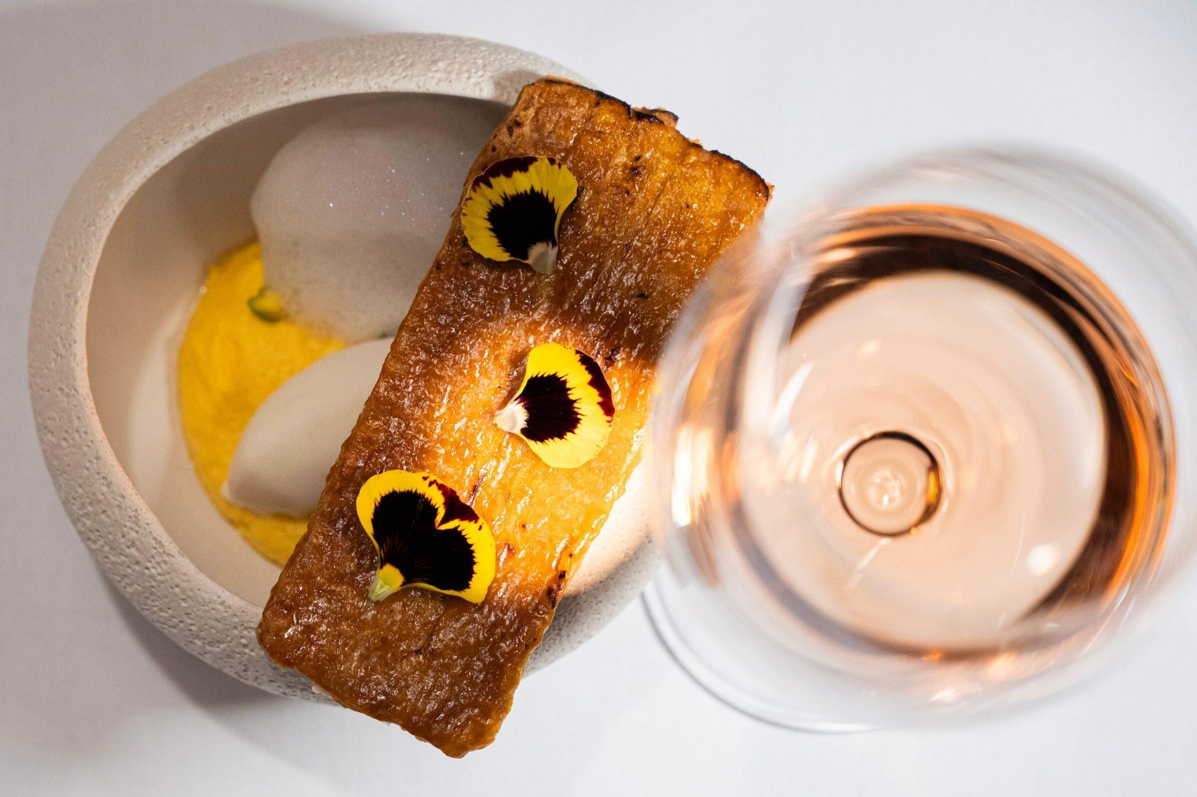 Ansicht von oben auf ein pflanzliches Gourmetgericht mit einem rechteckigen knusprigen Gebäck, das mit gelben und schwarzen Blüten garniert ist, neben einem Glas Roséwein auf einem weißen Tisch im veganen Hotel LA VIMEA in Südtirol.