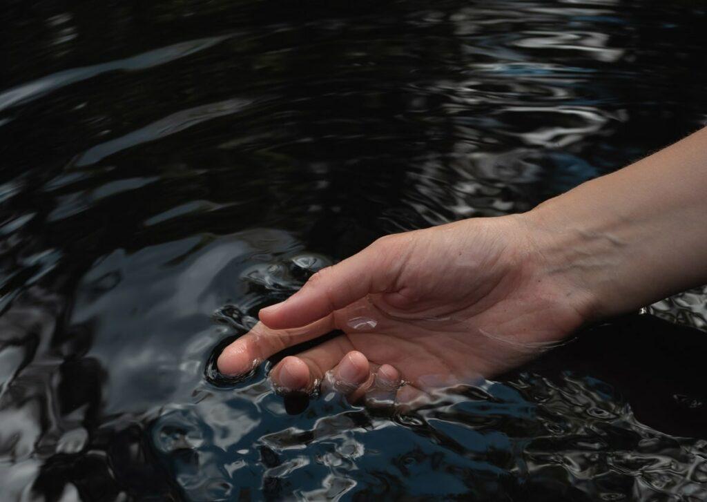 Eine Hand berührt sanft das plätschernde Wasser des Teiches im veganen Hotel LA VIMEA in Südtirol und erzeugt kleine Wellen. Die Szene vermittelt Gelassenheit und Ruhe, während Reflexionen auf der dunklen Wasseroberfläche tanzen.