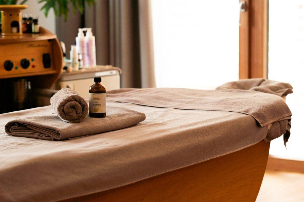 Ein gemütlicher Wellnessraum im Wellnesshotel LA VIMEA nur für Erwachsene mit einer mit beigen Handtüchern bedeckten Massageliege, einer Flasche Massageöl und einem aufgerollten Handtuch. Sanfte Beleuchtung schafft eine ruhige, entspannende Atmosphäre.