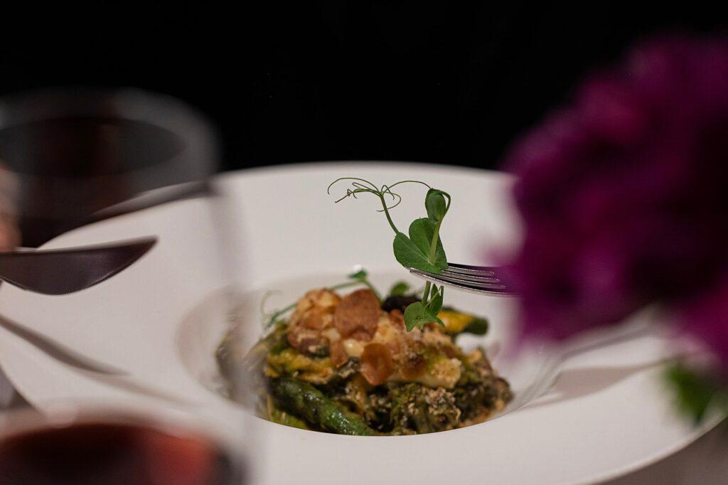 Pflanzliches Gourmetgericht im Restaurant LA VIMEA in Südtirol in einer weißen Schale, garniert mit zarten Erbsensprossen und knusprigen Toppings, umgeben von verschwommenen Weingläsern, die eine elegante Essatmosphäre schaffen.