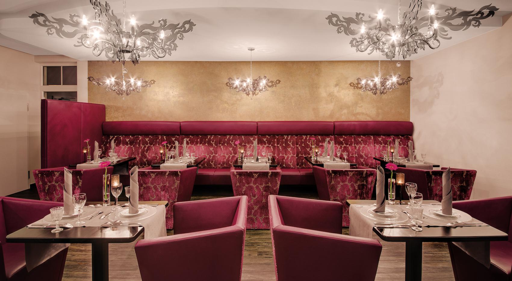 Restaurantbereich mit mehreren Tischen und Stühlen in dunklem Rot, dekorativen Kronleuchtern und einer goldfarbenen Wand im Lifestyle Resort Zum Kurfürsten in Bernkastel-Kues.