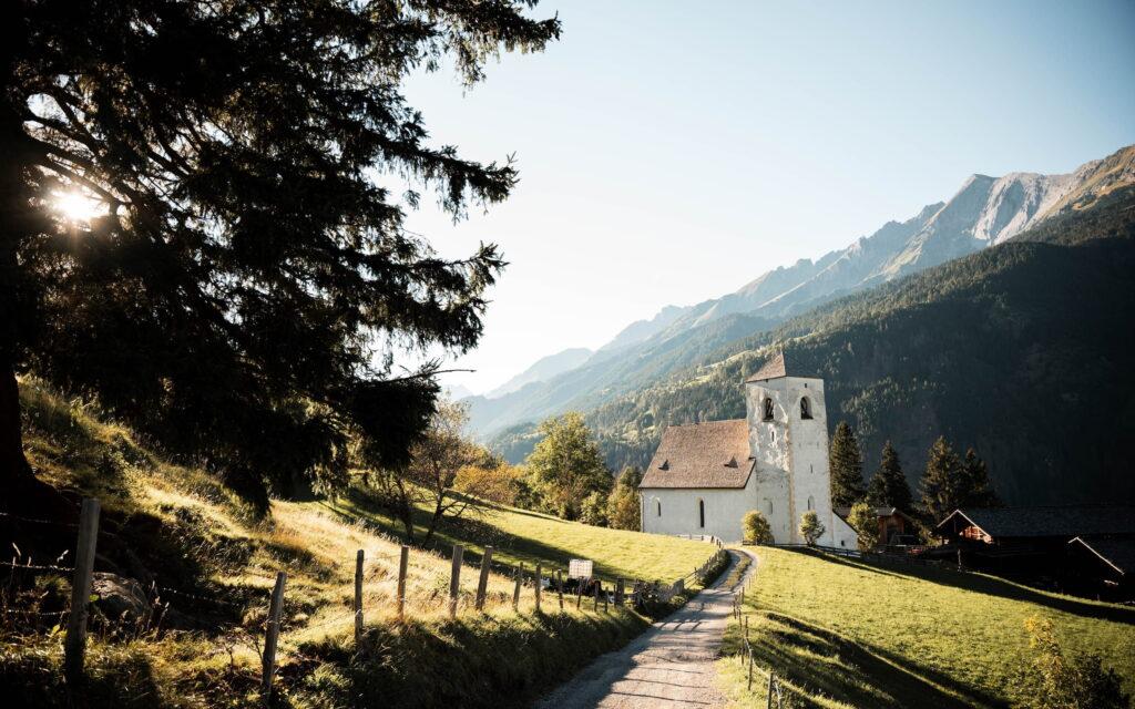 Eine ruhige Berglandschaft im österreichischen Nationalpark Hohe Tauern mit einem schmalen Feldweg, der zu einer malerischen weißen Kirche führt, umgeben von üppigem Grün, unter einem klaren blauen Himmel.
