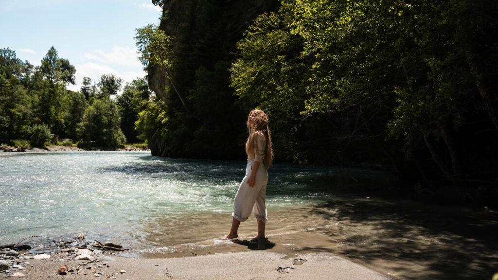 Eine Frau mit langen Haaren steht friedlich an einem sandigen Flussufer im Nationalpark Hohe Tauern, umgeben von üppigen, sonnenbeschienenen Bäumen. Die Szene ist ruhig und natürlich.