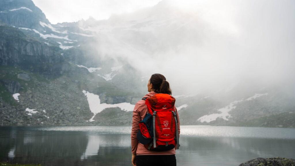 Eine Person mit einem roten Rucksack steht an einem nebligen Bergsee in Österreich, umgeben von felsigem Gelände und Schneeflecken, und vermittelt ein Gefühl von Abenteuer und Ruhe.