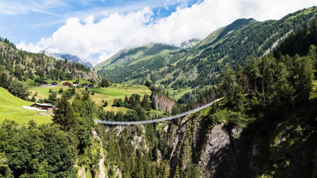 Eine schmale Hängebrücke überspannt ein grünes Tal im Nationalpark Hohe Tauern in Österreich, umgeben von hoch aufragenden Bergen und üppigen Wäldern unter einem strahlend blauen Himmel mit flauschigen Wolken.