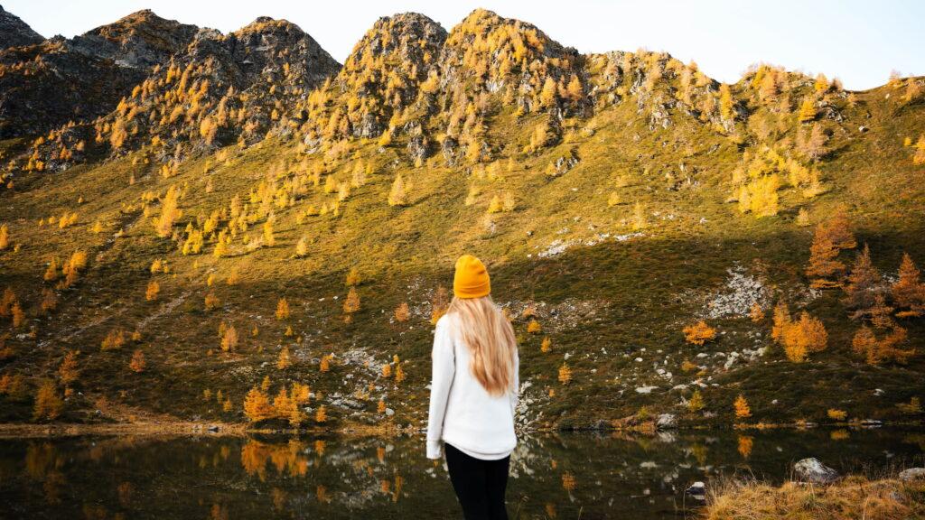 Eine Person mit langen blonden Haaren, einer weißen Jacke und einer gelben Mütze steht an einem ruhigen See und blickt unter einem klaren Himmel auf die sonnenbeschienenen, herbstlichen Alpen im Nationalpark Hohe Tauern, die Ruhe ausstrahlen.