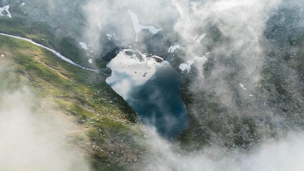 Luftaufnahme eines Bergsees im Nationalpark Hohe Tauern in Österreich, umgeben von Grasflächen und Felsen. Nebel und Wolken verdecken teilweise die ruhige Szenerie.