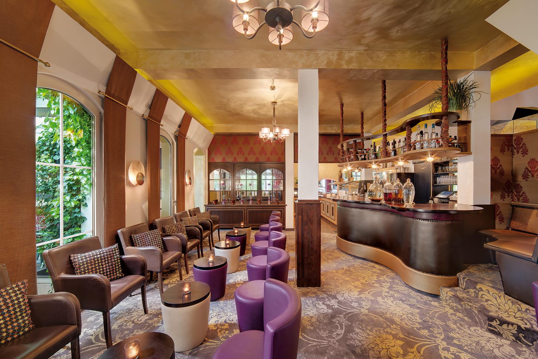 The bar area at the Zum Kurfürsten Lifestyle Resort features a curved counter, round purple stools, upholstered armchairs, and large windows overlooking lush greenery