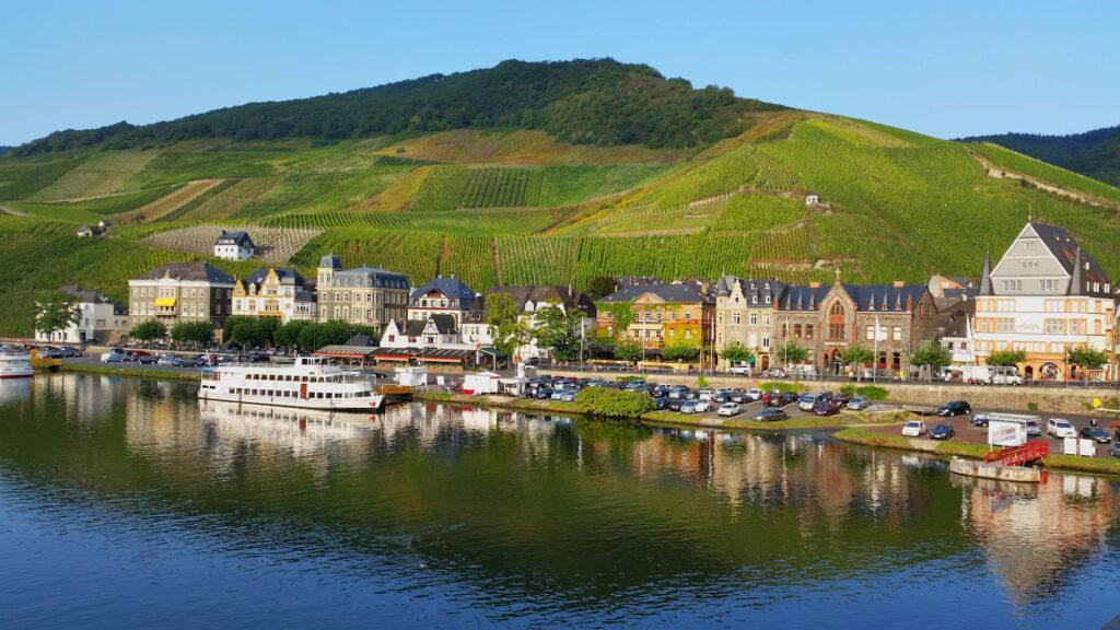 View of the Lifestyle Resort Zum Kurfürsten in Bernkastel-Kues, featuring the banks of the Moselle, a boat landing, parking lots, and vineyards in the background