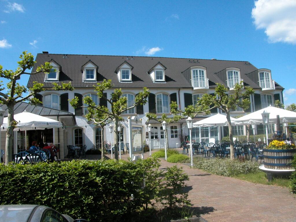 A multi-story building of the former Hotel Zum Kurfürsten, featuring a gabled roof and several dormers, set against a blue sky; in front of it, a terrace with tables, chairs, and umbrellas, as well as young trees and hedges