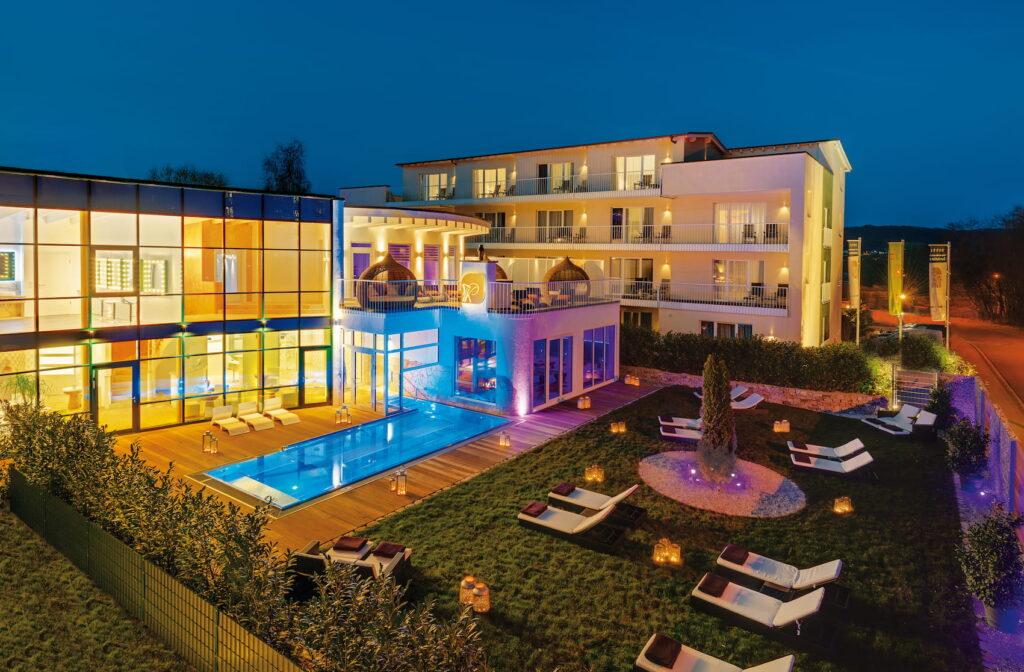The modern Zum Kurfürsten lifestyle resort on the Moselle, featuring an illuminated outdoor pool, lounge chairs on a well-manicured lawn, and a multi-story building bathed in the evening light